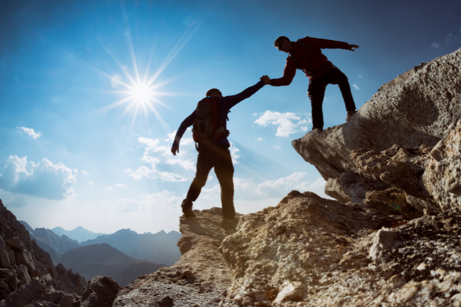 Zwei Menschen beim Bergsteigen, der Eine hilf dem Anderen hoch auf den Berg. Symbolbild für das Erreichen eines MBA Titels, welcher durch Hilfe und Unterstützung des richtigen Anbieters möglich ist.