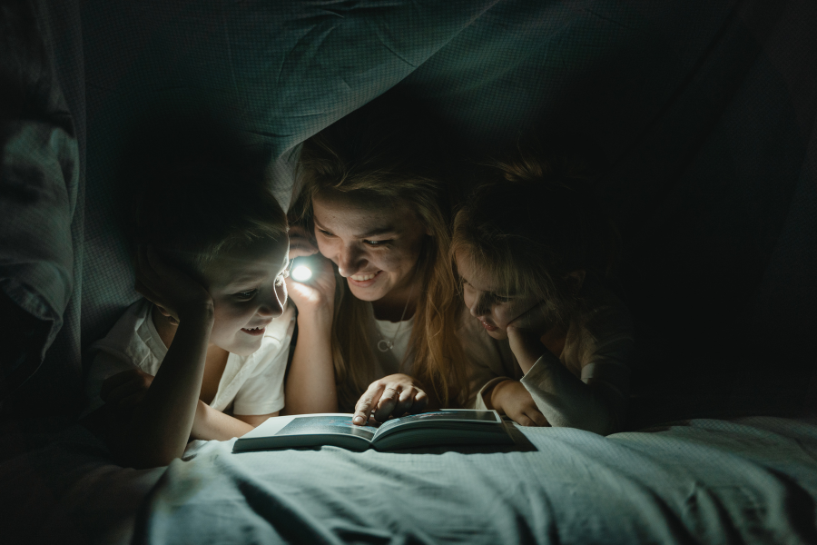 Frau mit Kindern, Taschenlampe und Buch unter Bettdecke beim Geschichten erzählen. Symbolbild für Storytelling Online Kurs für Influencer und KMU.